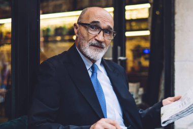 Gazete ve bardaklı kahve fincanı ve takım elbiseli yaşlı sakallı bir erkek sokakta oturup pencerelere bakıyor.