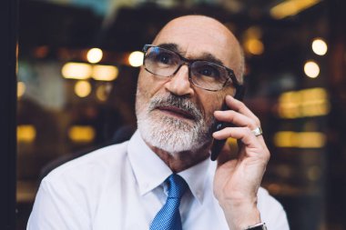 Gözlüklü, takım elbiseli, beyaz gömlekli, akıllı telefondan konuşan, camlara bakan yaşlı, kısa saçlı, yakışıklı bir erkek.