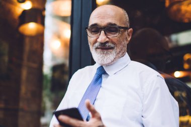 Yaşlı, kısa saçlı, gözlüklü, beyaz gömlekli, sokak kafesinin penceresinde oturan ve akıllı telefondan mesaj atan bir erkek.