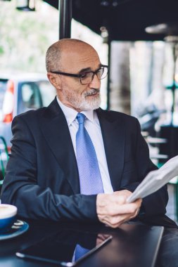 Ofis kıyafeti giyen, kravat takan ve gözlüklü neşeli, son sınıf kel bir adam sokak kafesinde oturmuş, bulanık şehir geçmişi hakkında gazete okuyor.