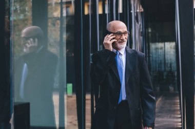 Resmi kıyafetli, gözlüklü, beyaz sakallı, sakallı, sakallı yaşlı bir erkek sokakta yürürken telefonla konuşuyor.