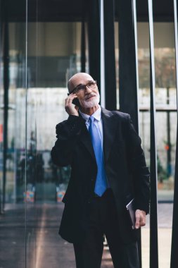 Takım elbiseli, gözlüklü, beyaz sakallı, tabletli yakışıklı bir erkek sokakta yürürken telefonla konuşuyor.