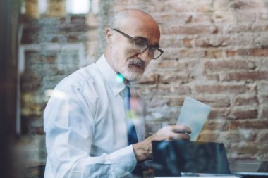 Gözlüklü, yaşlı, kel bir adamın resmi kıyafetlerini incele. Kafede dizüstü bilgisayarla otururken not al.