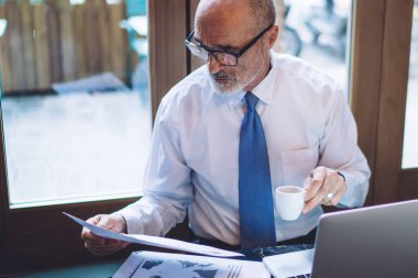 Yukarıdan bakıldığında, gözlüklü, elinde bir fincan kahve kağıdıyla masada, pencerenin yanında dizüstü bilgisayarlı bir şekilde otururken görülüyor.