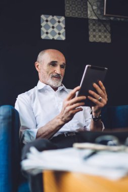 Beyaz gömlekli, siyah pantolonlu, kel, yaşlı bir adam kafede otururken tablet kullanıyor.