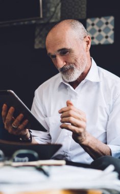 Beyaz tişörtlü, beyaz sakallı, kel bir adam oturma odasında otururken tablet ekrana bakıyor.