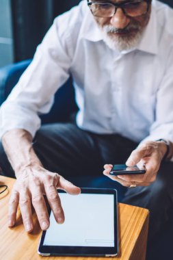 Resmi kıyafetler ve gözlüklü yaşlı adam rahat koltukta oturuyor, akıllı telefona bakıyor ve boş tablet ekranına dokunuyor.