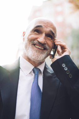 Resmi kıyafetli, yakışıklı, yaşlı ve beyaz sakallı bir erkek sokakta yürürken telefonla konuşuyor. Bulanık binaların arka planına bakıyor.