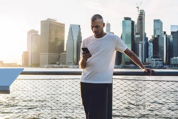 Kısa saçlı, spor elbiseli, gökdelenlerin arka planında duran ve akıllı telefondan ciddi bir yüzle mesaj atan genç bir adam.