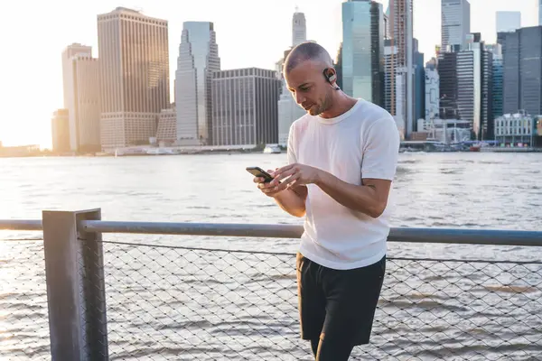Spor giyim sektöründe genç ve yakışıklı bir erkek modern akıllı telefonda kulaklık takarken nehir kenarında gökdelenlerle dolu bir arka planda kulaklık takıyor.