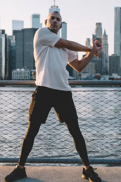 Spor giyim ve kulaklıklarda genç ve çekici kel bir adam koşmadan önce ısınıyor ve gökdelenlerle dolu kentsel arka plandaki sete bakıyor.