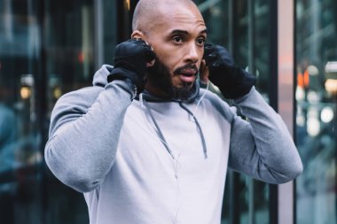 Spor kıyafetli kel Afrikalı Amerikalı adam modern ofis binasının girişinde dikilirken kulaklık takıyor.