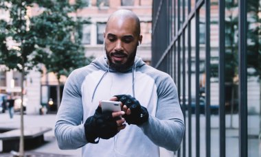 Atletik kel sakallı Afro-Amerikan sporcu kulaklıkla müzik dinliyor ve modern ofis binasının yanındaki kaldırımda dikilirken akıllı telefonuna bakıyor.