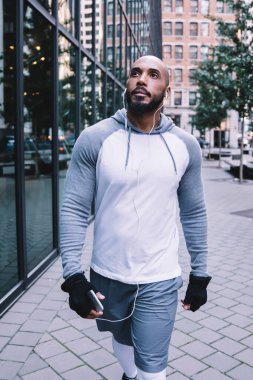 Spor kıyafetlerine güveni tam atletik kel bir adam elinde akıllı telefonuyla kulaklıklarla sokakta yürüyor ve gözlerini kaçırıyor.