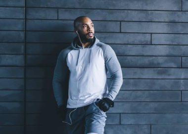 Beyaz kulaklıklı gri spor giysili, kel, atletik bir sporcu. Bacağı duvara dayanmış, başka tarafa bakarken müzik dinliyor.