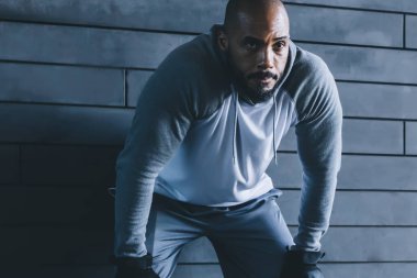 Gri spor giyimli, kel ve yorgun bir adam duvara yaslanmış, dizlerinin üstüne çökmüş ve antrenman sırasında fotoğraf makinesiyle ekin fotoğrafçısına bakıyor.