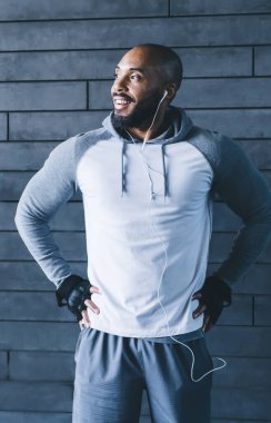 Beyaz kulaklıklı gri spor giysili, kaslı, kel sporcu. Kalçasında beyaz kulaklıklar var ve taş duvarın yanından başka yere bakıyor.