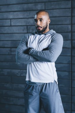 Yakışıklı, ciddi ciddi atletik Afrikalı, kel, sakallı, spor kıyafetli, kollarını çapraz yapıp, gri arka plana bakan yetişkin bir erkek.