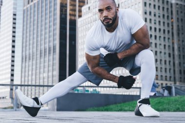 Spor giyimli kel sakallı Afrikalı Amerikalı adam modern şehir merkezinin sokaklarında antrenman yapmadan önce yan atlama ve esneme hareketleri yapıyor.