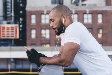 Spor kıyafetleri içinde siyah sakallı kel bir erkek akıllı telefondan mesaj atıyor ve şehir merkezindeki ahşap iskelede korkuluklara yaslanırken müzik dinliyor.