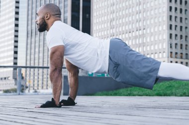 Kel sakallı Afro-Amerikan sporcu spor kıyafetleri içinde modern şehir merkezinin sokaklarında arka planda gökdelenler eşliğinde tahta gösterisi yapıyor.