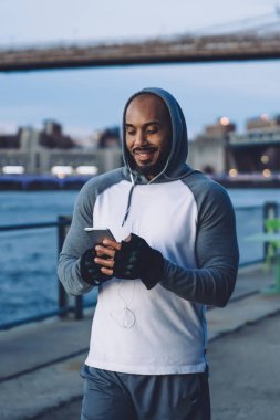 Modern spor giyim ve kulaklıklarda akşamları Megapolis rıhtımında bulanık arka planda dururken cep telefonu kullanan güçlü siyah adam.