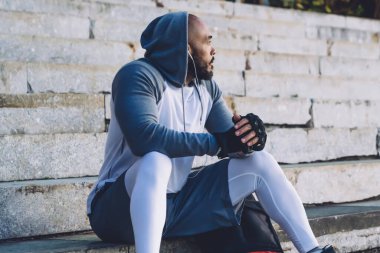 Genç Afrikalı Amerikalı sporcu sırt çantasının yanındaki taş merdivenlerde oturuyor ve kulaklıkla müzik dinliyor.