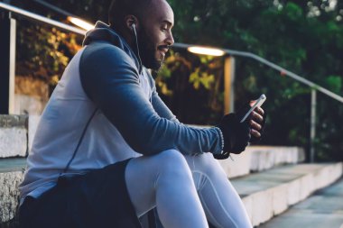 Spor kıyafetleriyle merdivenlerde oturan ve kulaklıkla müzik dinleyen akıllı telefondan mesaj atan konsantre Afro-Amerikan genç adamın yan görüntüsü.