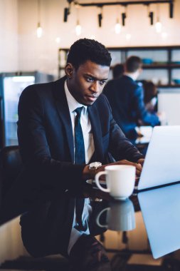 Ciddi bir Afro-Amerikan erkek ofis çalışanı ofis dinlenme alanında kahve içerken dizüstü bilgisayarla proje üzerinde çalışıyor.