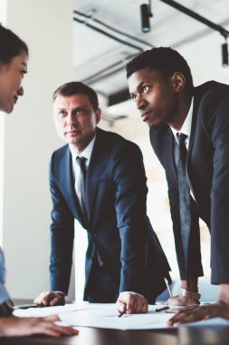 Çok ırklı genç iş arkadaşları ellerinde cihazlar ve belgelerle masanın yanında duruyor ve proje planını şirket ofisindeki toplantı odasında analiz ediyorlar.