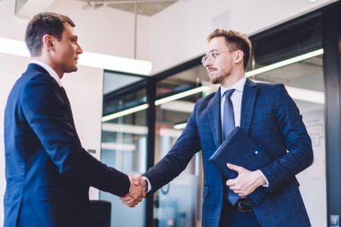 Modern ofiste giyinmiş iki kendine güvenen iş adamı buluşmayı bitiriyor ve birbirlerine bakarken tokalaşıyorlar.