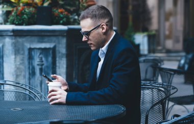 Takım elbiseli ve gözlüklü bir gencin Cafe 'deki masada oturup telefon ve kahve içerkenki görüntüsü.