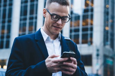 Siyah ceketli genç adam modern ofis binasının dışındaki caddede duruyor ve bulanık arka planda cep telefonuyla mesajlaşıyor.