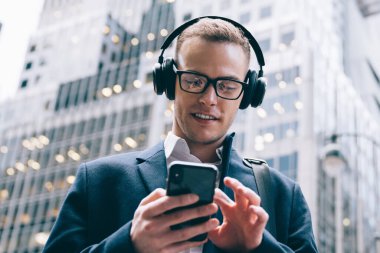 Gözlüklü, resmi ve yakışıklı bir erkek çalışandan kablosuz kulaklıkla müzik dinlerken ticari binaların bulanık arka planında cep telefonuna göz gezdiriliyor.