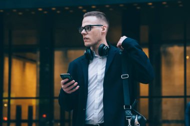 Kendine güveni tam genç girişimci resmi giyinmiş ve gözlüklü akıllı telefon kullanarak modern ofis binasının önünde bulanık bir şekilde yürüyordu.