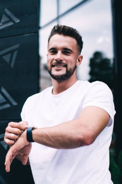 Beyaz tişörtlü mutlu erkek modern akıllı saati kontrol ediyor ve ofis binasının yakınında beklerken kameraya bakıyor.