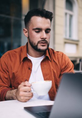 Turuncu tişörtlü, uzaktan kumandalı iş yaparken dizüstü bilgisayar izleyen ve kahve içen kendine güvenen genç bir adam.