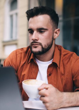 Turuncu tişörtlü, ciddi yakışıklı bir adam. Laptopunu karıştırıyor ve yazın kafede kahve içiyor. Masada uzaktan çalışıyor.