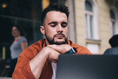 Düşünceli, sakin, yakışıklı, elleri çenesine dayanmış ve yazlık kafede kahve içmeye giderken dizüstü bilgisayarında içeriğini izleyen bir erkek.