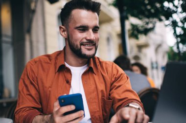 Gülümseyen turuncu tişörtlü, modern dizüstü bilgisayarını karıştıran ve elinde cep telefonu tutan genç bir erkek yazlık kahve evinde.