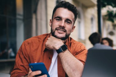 Turuncu tişörtlü neşeli yakışıklı adam dışarıda laptopuyla kafeteryada otururken kameraya bakıp telefonla mesaj atıyor ve çenesine dokunuyor.