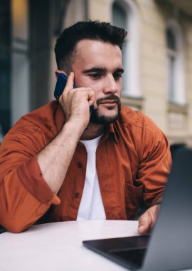 Kafedeki masada dizüstü bilgisayar kullanırken endişeyle akıllı telefondan konuşan serbest giyinmiş dalgın genç bir erkek.