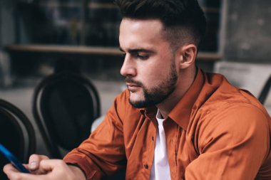 Düşünceli genç erkek müşteri kafeteryada dizüstü bilgisayarla otururken dikkat ve ilgi ile akıllı telefona göz atıyor.