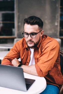 Düşünceli, genç, gözlüklü, günlük kıyafetler giyen, kafedeki masada otururken dikkatle dizüstü bilgisayara bakan serbest bir erkek.