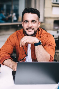 İyimser erkek öğrenci, gündelik kıyafetlerle, akıllı saatle bilgisayarda çalışıyor ve kafede kahve içerken kameraya gülümsüyor.