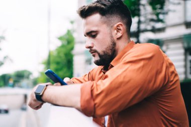 Hippi görünümlü, sakallı, sıradan kıyafetli, akıllı telefonlu, sokakta çitlere yaslanmış bir adam.