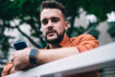 Aşağıdaki hippi görünümlü sakallı sıradan turuncu tişörtlü adamdan. Elinde akıllı bir telefon, çitlere yaslanmış, caddede duruyor ve kameraya bakıyor.