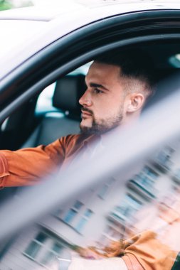 Sıradan giysiler içinde konsantre olmuş sakallı bir erkek sürücü elleri direksiyonda oturmuş camları açık, dikkatle başka tarafa bakıyor.