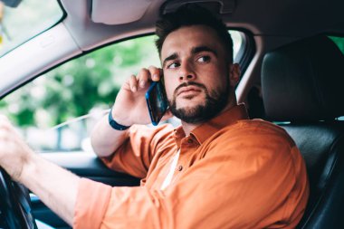 Pahalı bir arabada oturan, direksiyonu çeviren, akıllı telefondan konuşan ve arka planda bulanık duran erkek sürücünün yan görüntüsü.