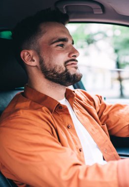 Turuncu tişörtlü, sakallı, gülümseyen ve direksiyon sallayan, şehrin bulanık arka planında...